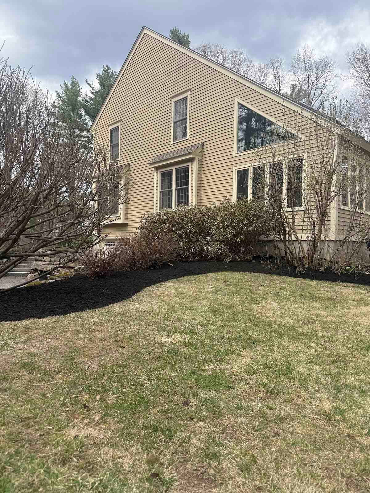 Tan house with steep roof, windows, and shrubs on a grassy hill under cloudy trees