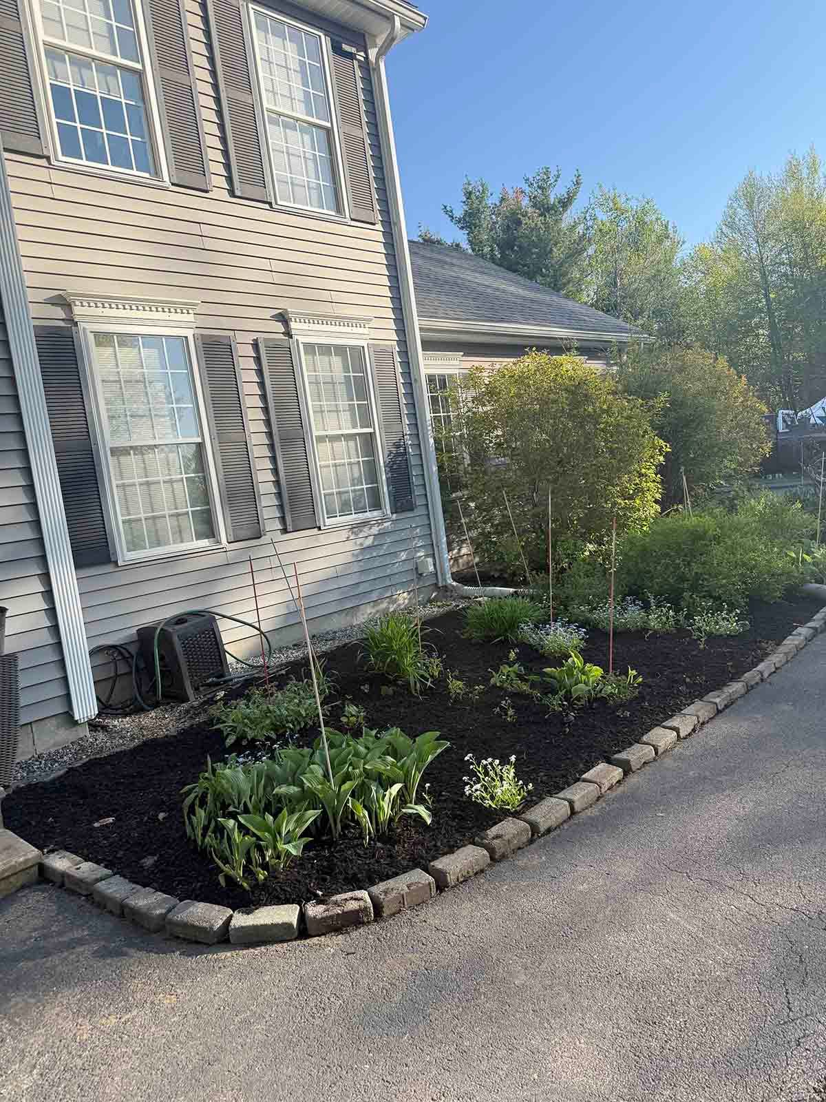 Front of a house with a landscaped garden bed, black mulch, shrubs, and stone edging along a driveway