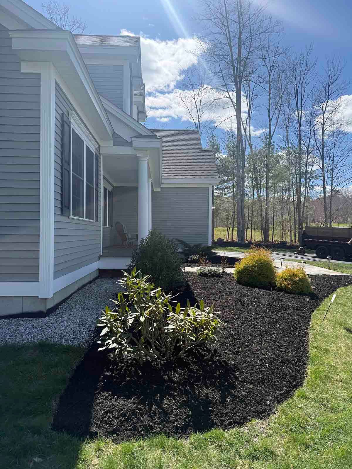 Side view of a gray house with white trim, landscaped shrubs, and a sunny blue sky