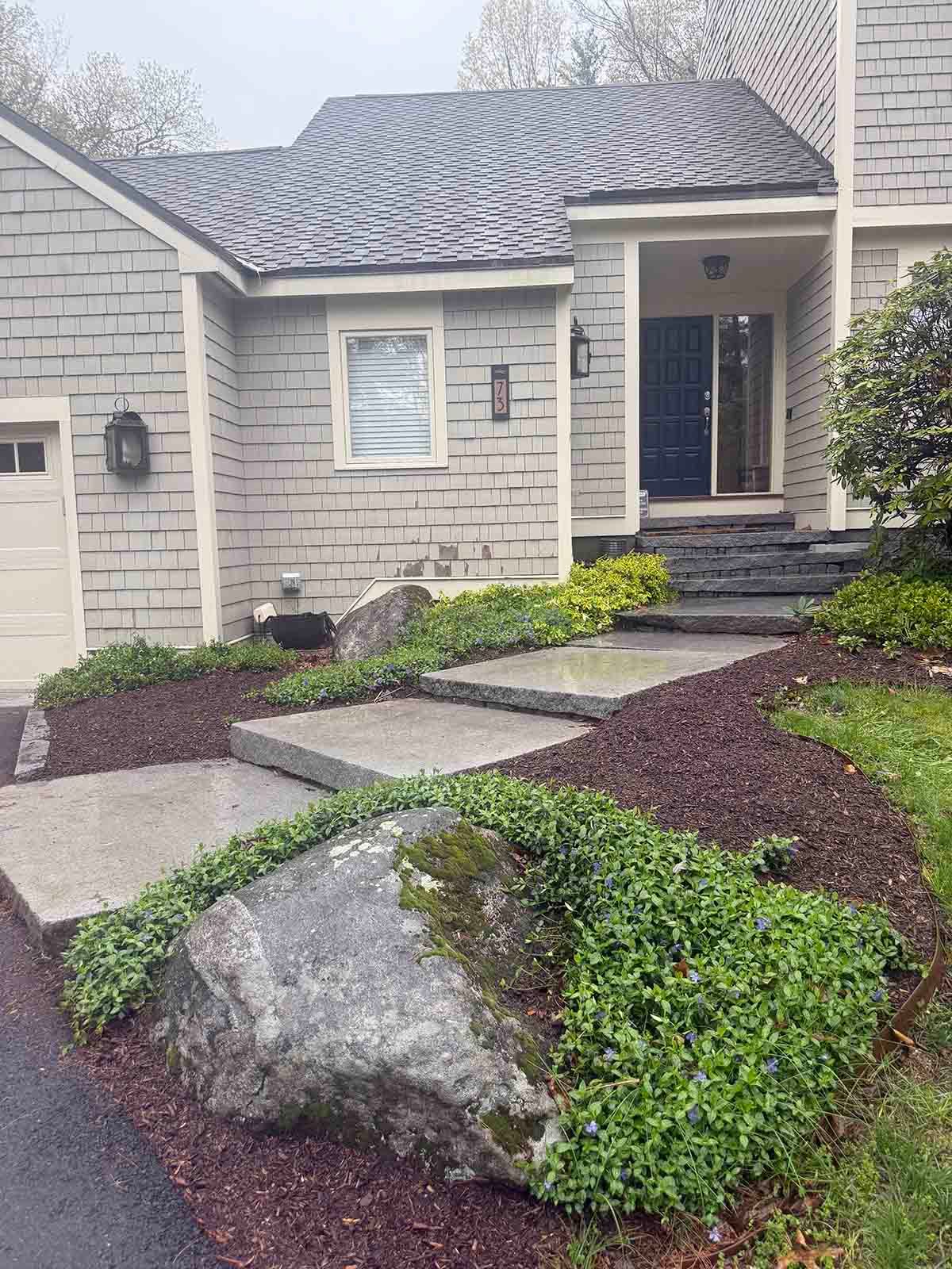 Front walkway to a stone house with steps, landscaping, and a large boulder in the foreground
