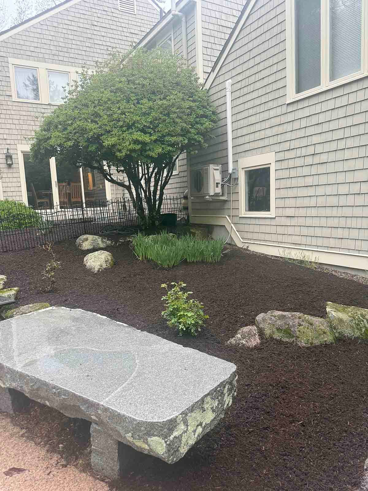 Landscaped courtyard with shrubs, rocks, and a stone bench beside a gray house