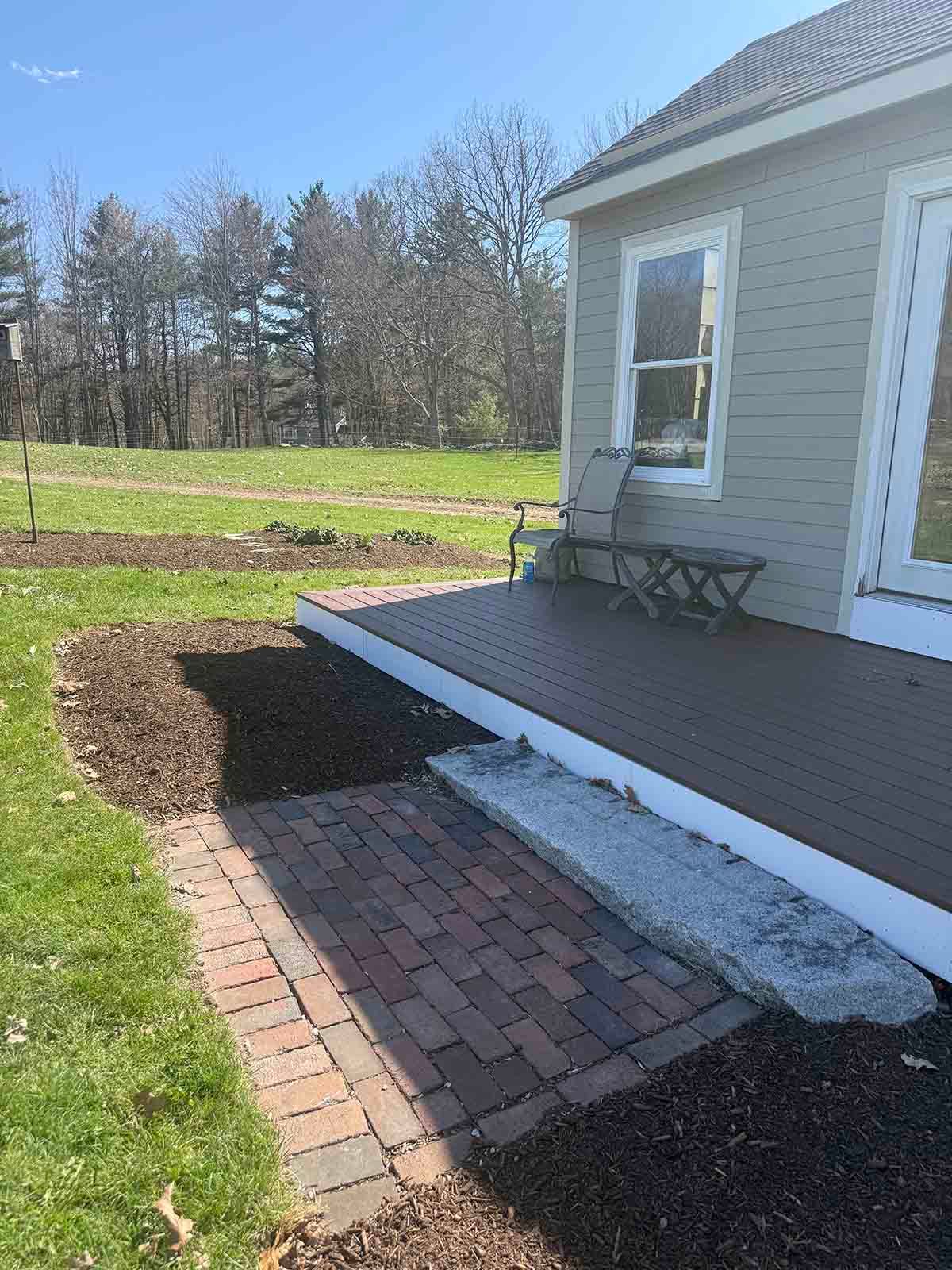 Brick walkway beside a raised porch with a white house, small steps, and lawn on a sunny day