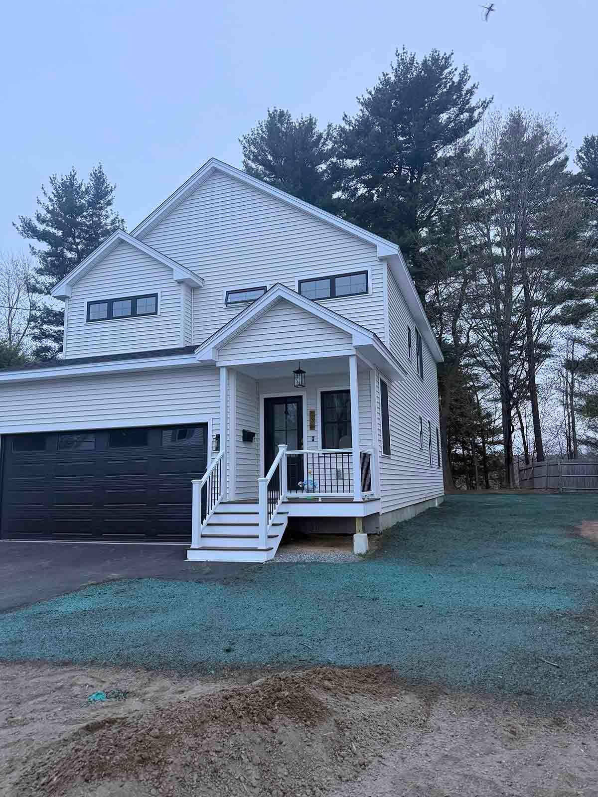 White two-story house with black garage, front porch, and snow-dusted yard among tall trees.