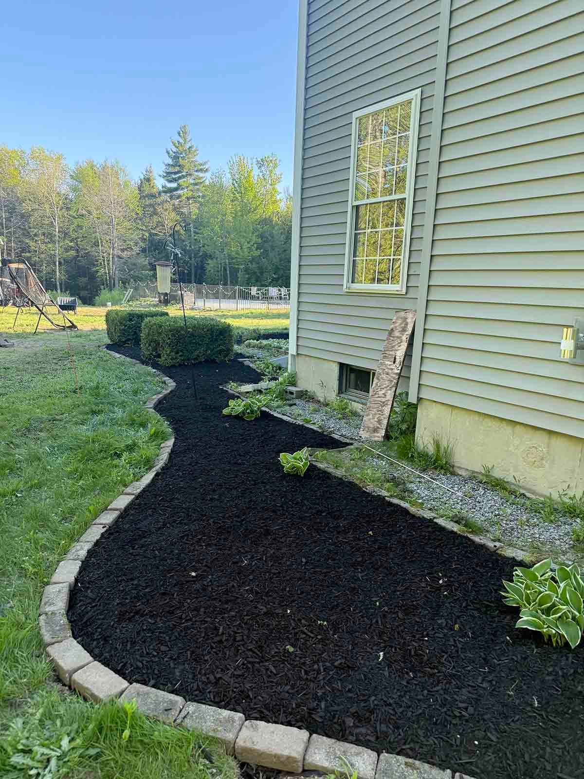 Curved stone-edged mulch bed beside a house with gray siding, near a window and lawn.
