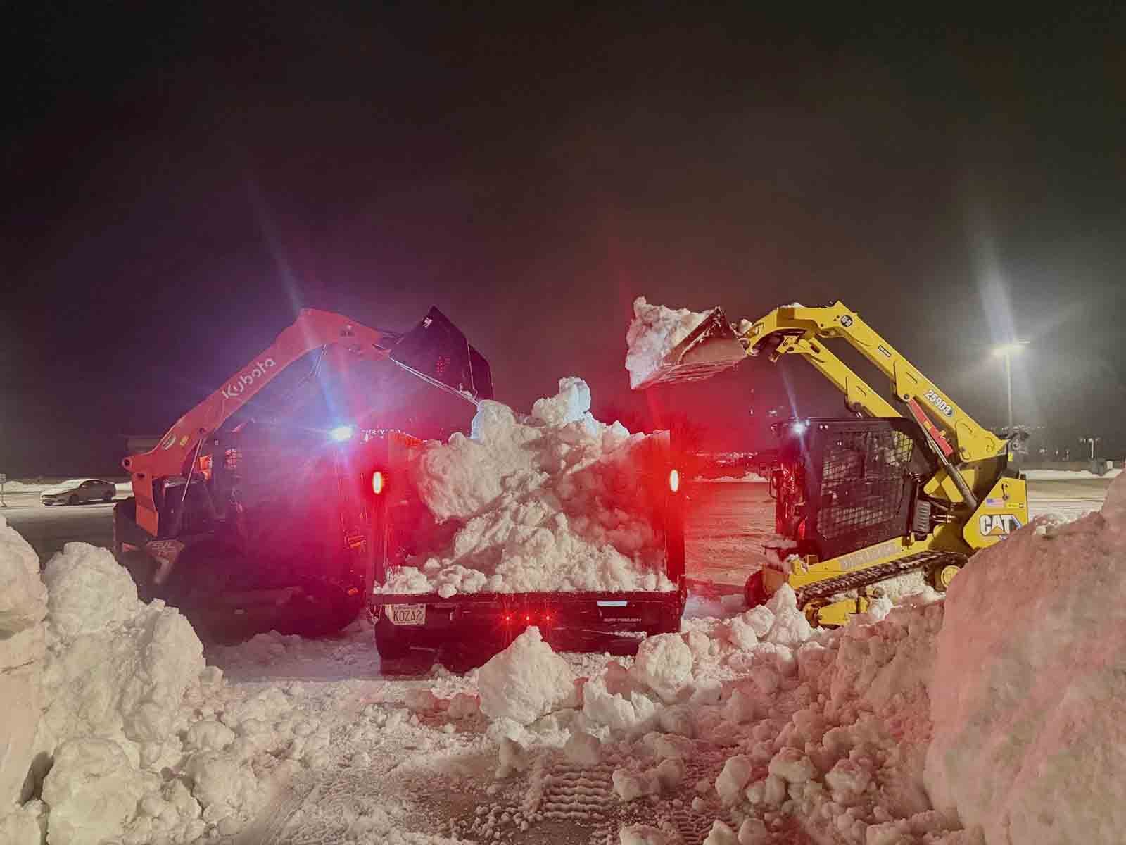 Two snowplows clearing a snowy road at night with bright red and yellow lights