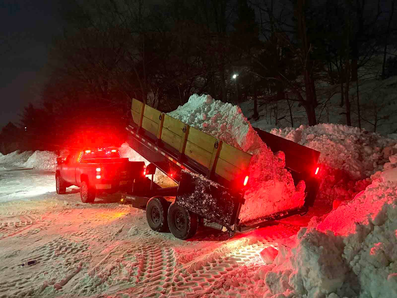 Tow truck hauling a dump trailer through snow at night, red lights glowing as it climbs a snowy road