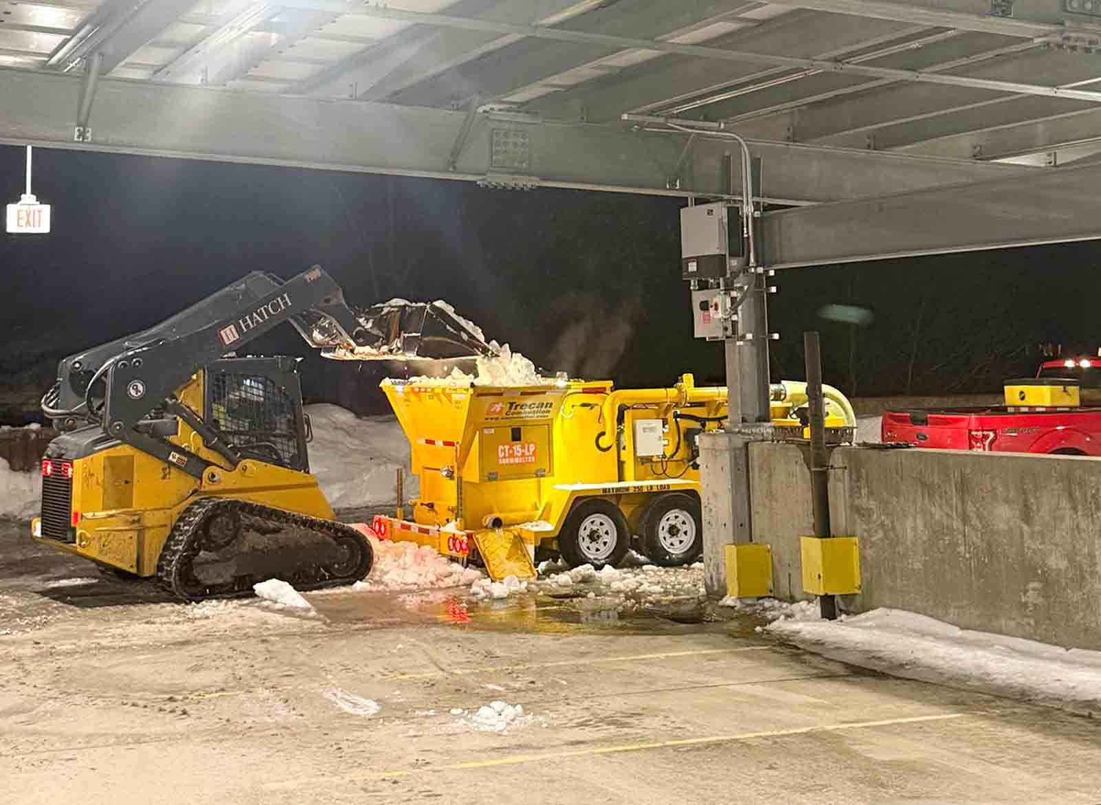 Yellow loader dumping snow into a yellow dump truck at a snowy work site under a covered structure.