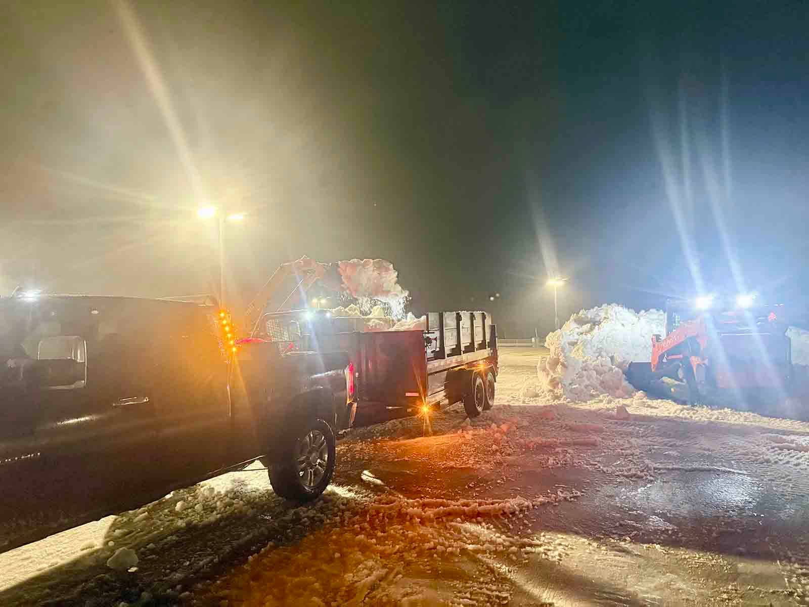 Snowplow and trucks clearing a snowy road at night under bright headlights and streetlights