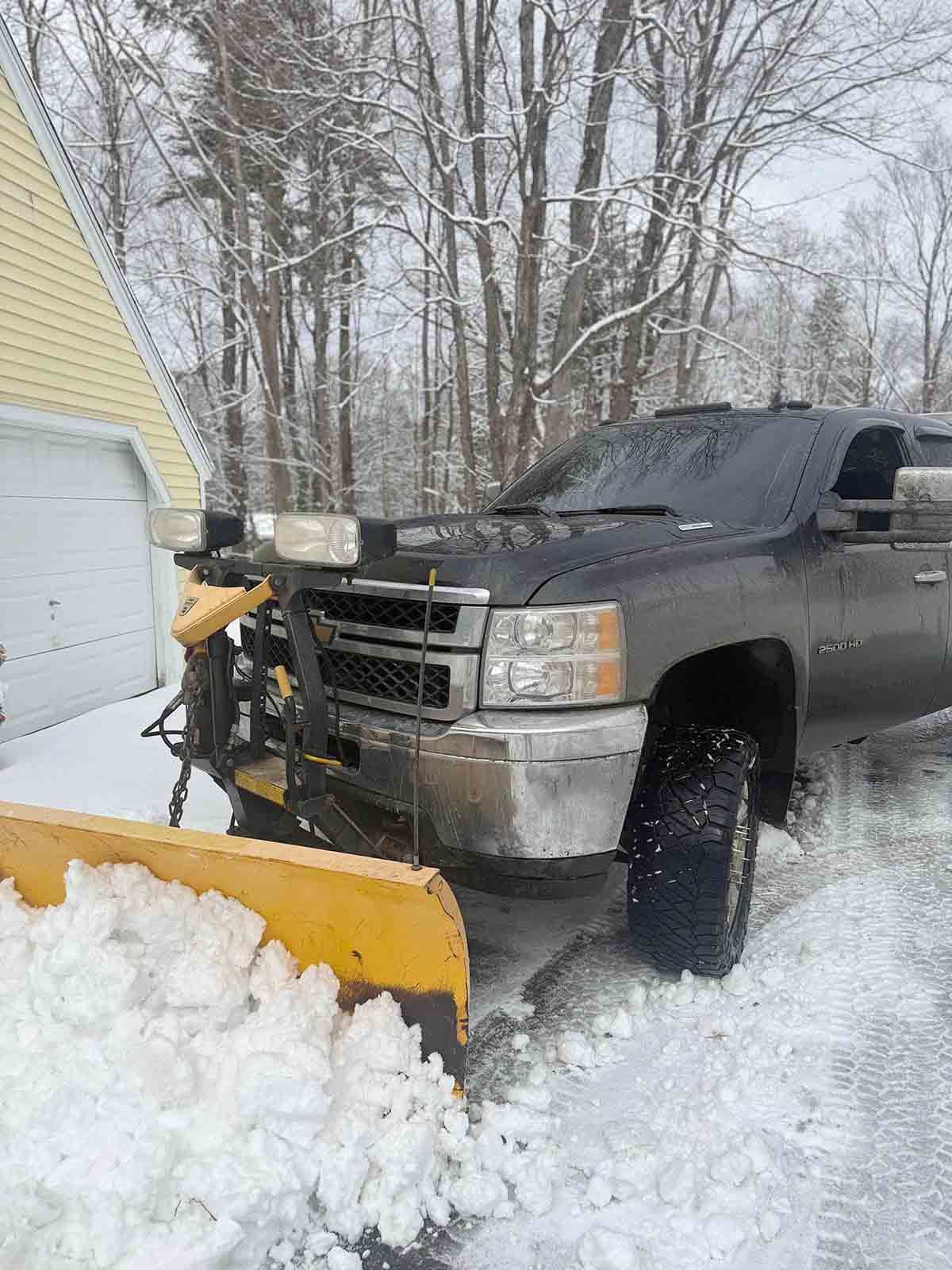 Black pickup truck with snow plow clearing a snowy driveway by a garage in winter