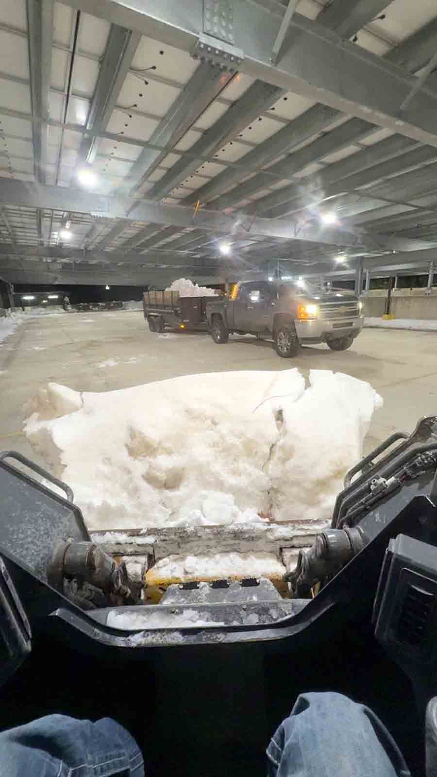 Inside a warehouse, a snowplow clears snow while viewed from a vehicle cabin.