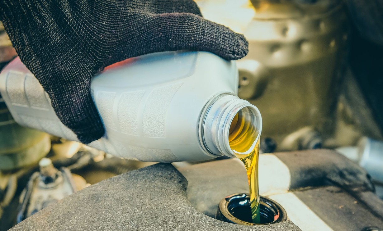 Gloved hand pouring oil from a white bottle into a car engine.
