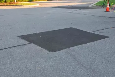 Dark square patch on a paved road near a curb and traffic cones.