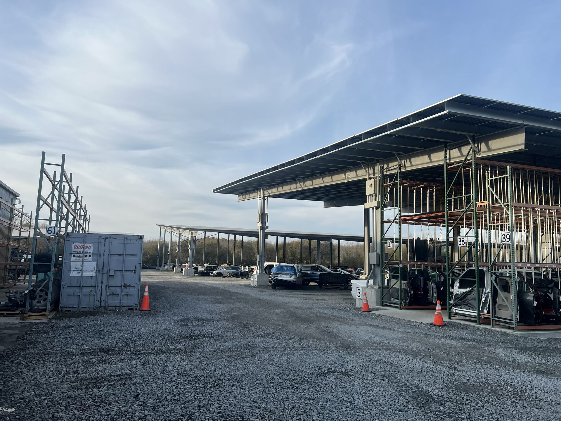 Gravel lot with rows of parked cars under canopies, storage racks on the right, and a shipping container on the left, under a cloudy sky.