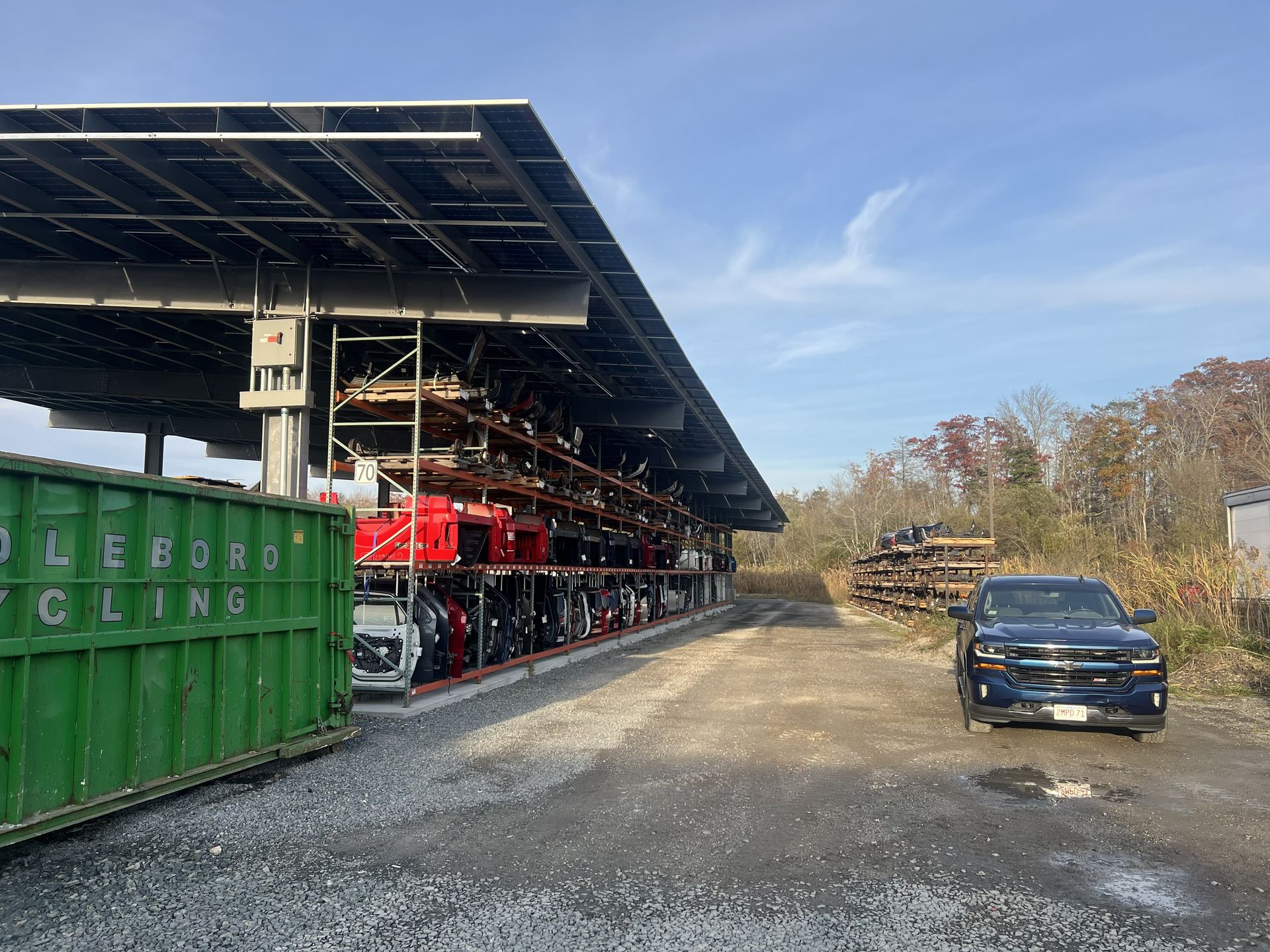 Solar panel covered storage racks filled with auto parts next to a recycling dumpster and truck.