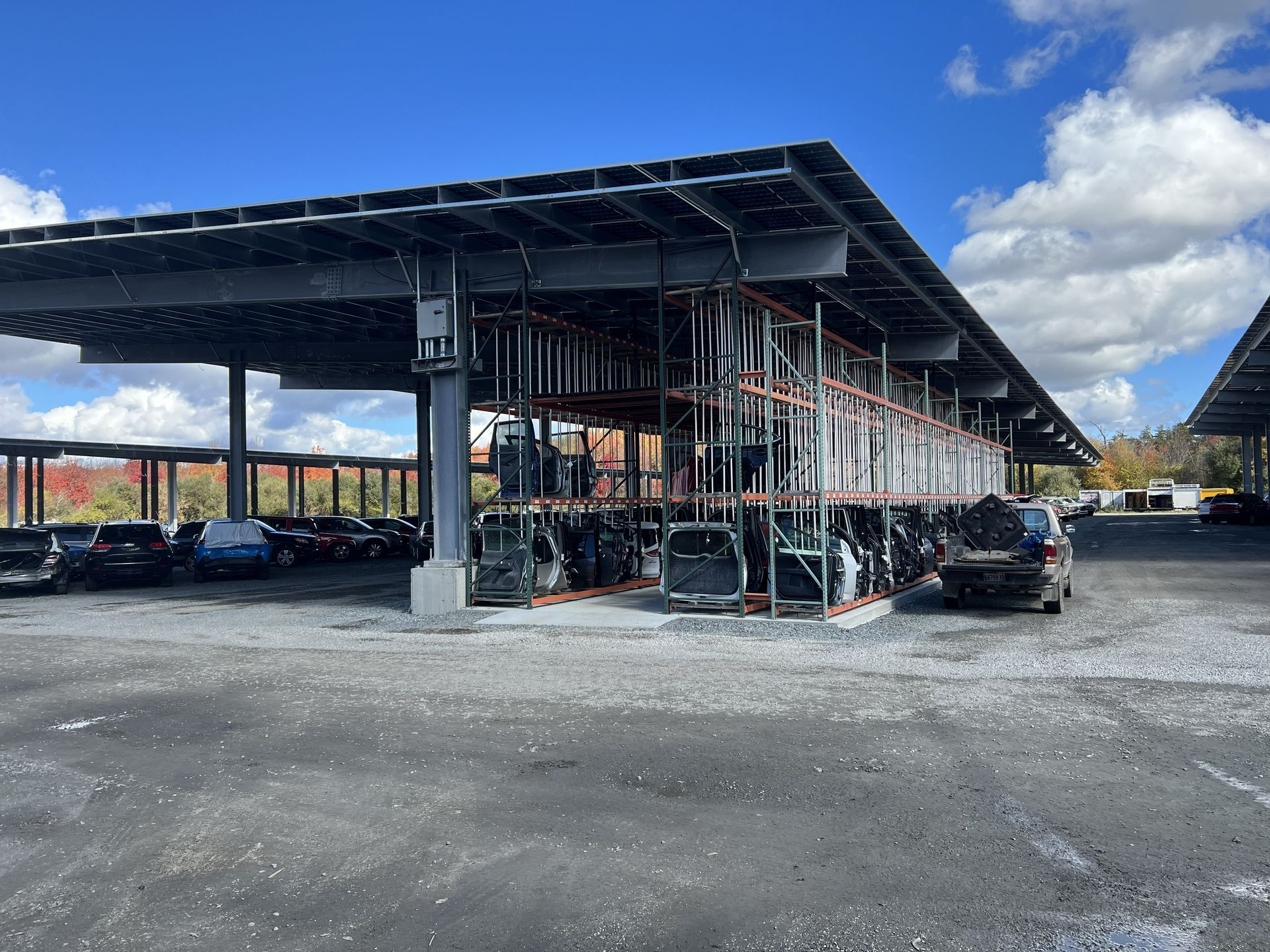 Car parts storage under a metal shelter with parked cars in a salvage yard on a bright day.