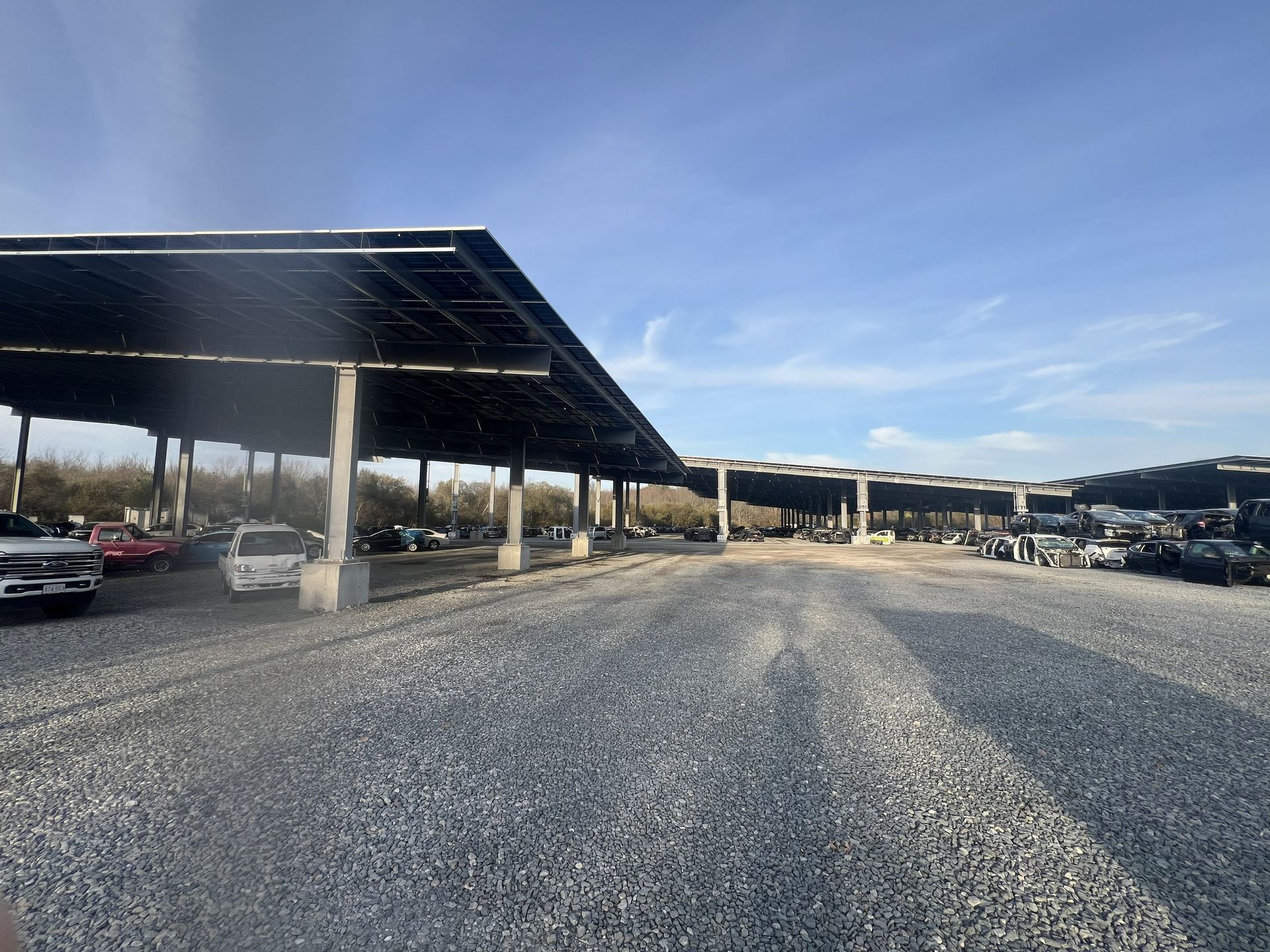 A solar panel-covered parking lot with gravel ground, cars parked under the panels, and a blue sky.