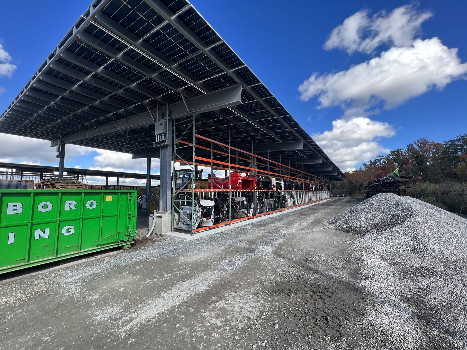 Solar panel canopy over a gravel lot with various vehicles parked beneath, bright sky above.
