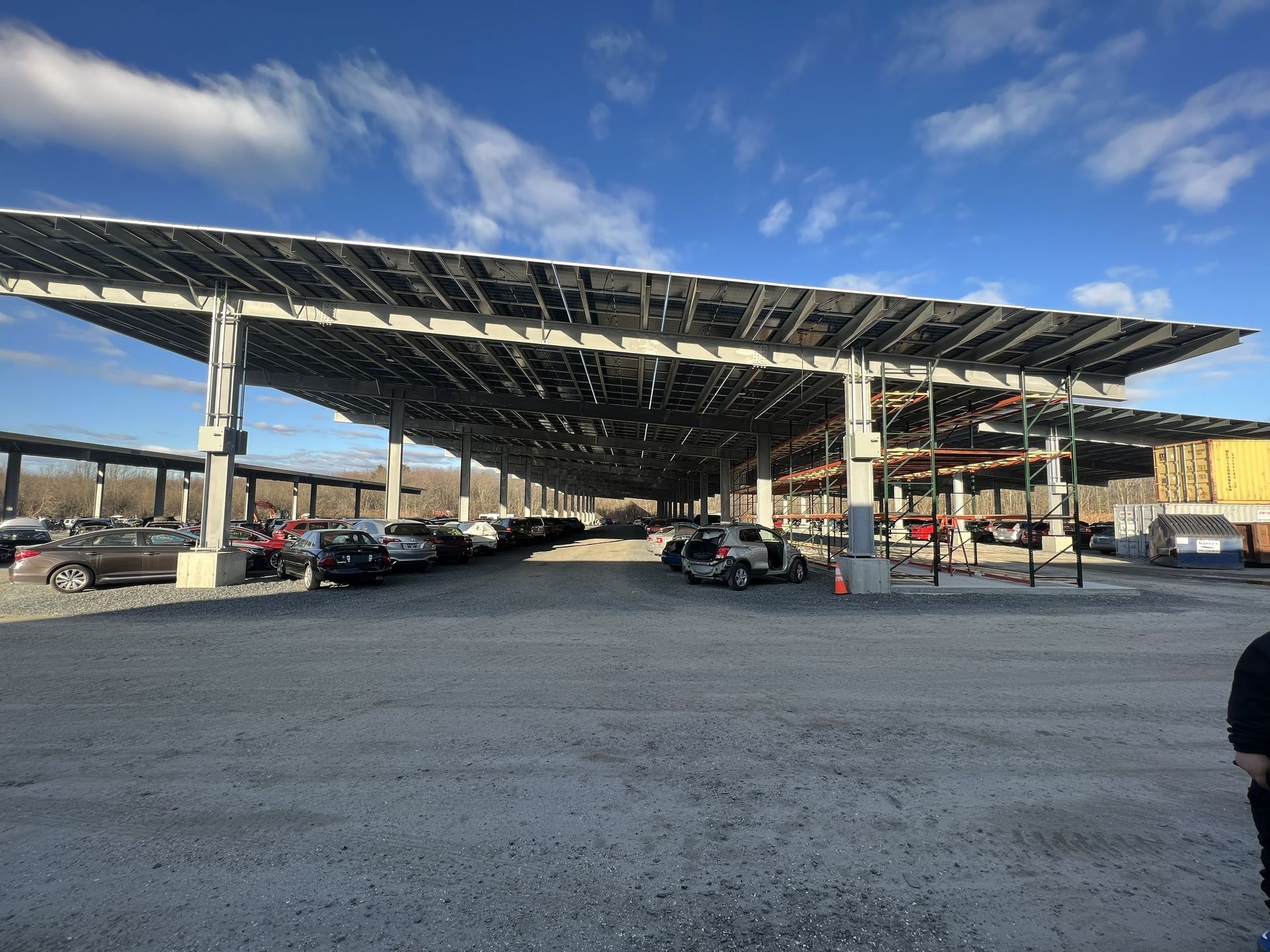 Solar panel carport over parked cars on a gravel lot under a partly cloudy blue sky.