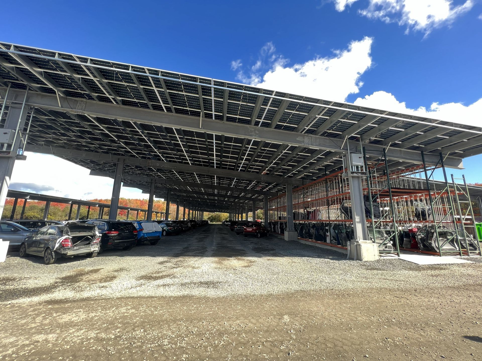 Solar panel covered parking lot, cars parked beneath. Gravel ground, sunny day.