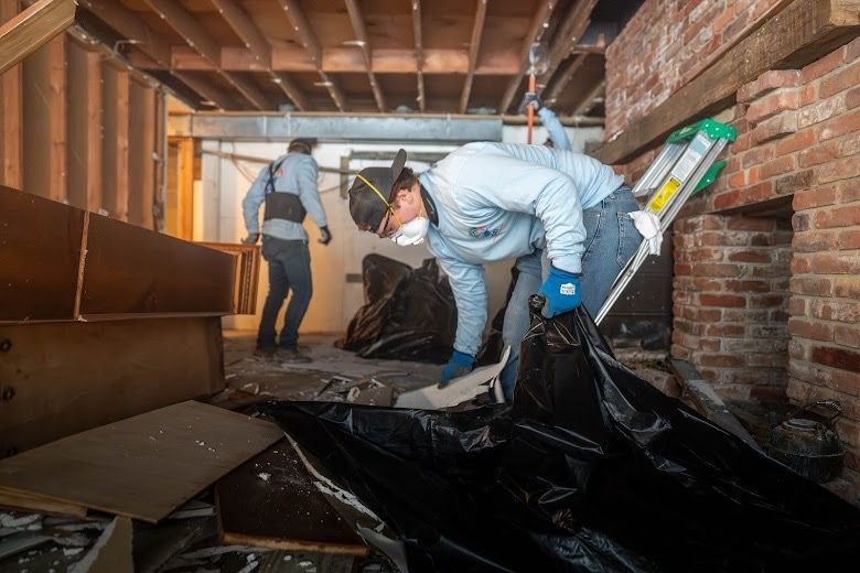 Workers in a crawlspace clearing debris under exposed floor joists and brick walls
