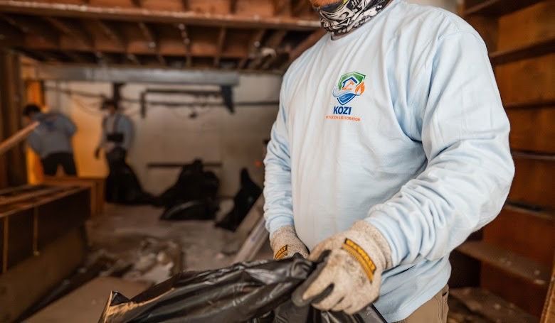 Worker in white uniform and gloves sorting black trash bags in a warehouse-like room.