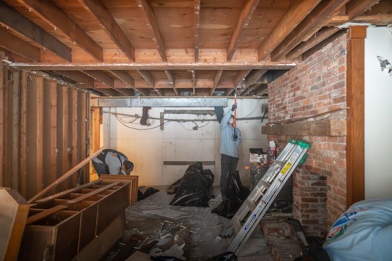 Unfinished basement with exposed wood beams, brick wall, tools, and debris during renovation