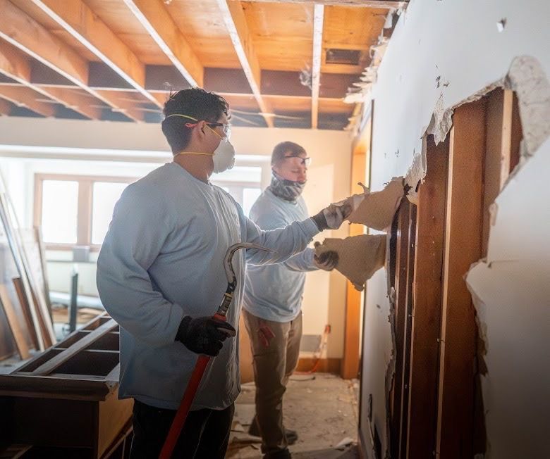 Two workers demolishing a wall in a partially renovated room with exposed beams and insulation.