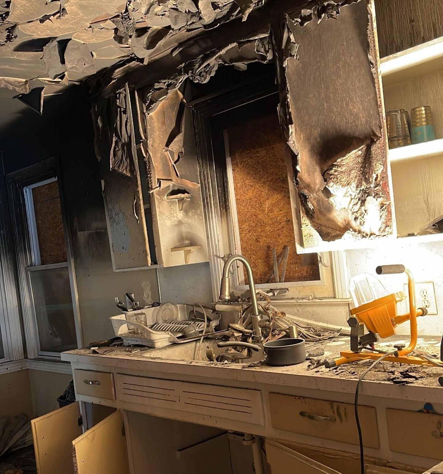 Fire-damaged kitchen with charred cabinets, collapsed ceiling, and debris on the counter.