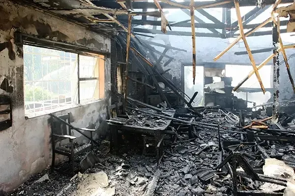 Fire-damaged room with charred debris, collapsed roof beams, and a smoke-blackened interior