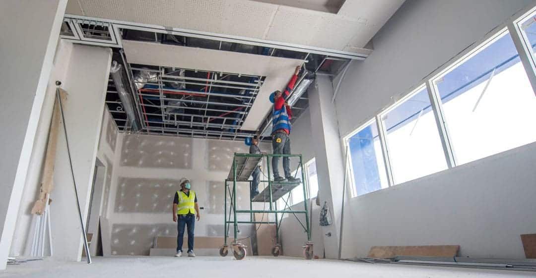 Workers installing a ceiling in a bright room under renovation, with scaffolding and exposed wiring
