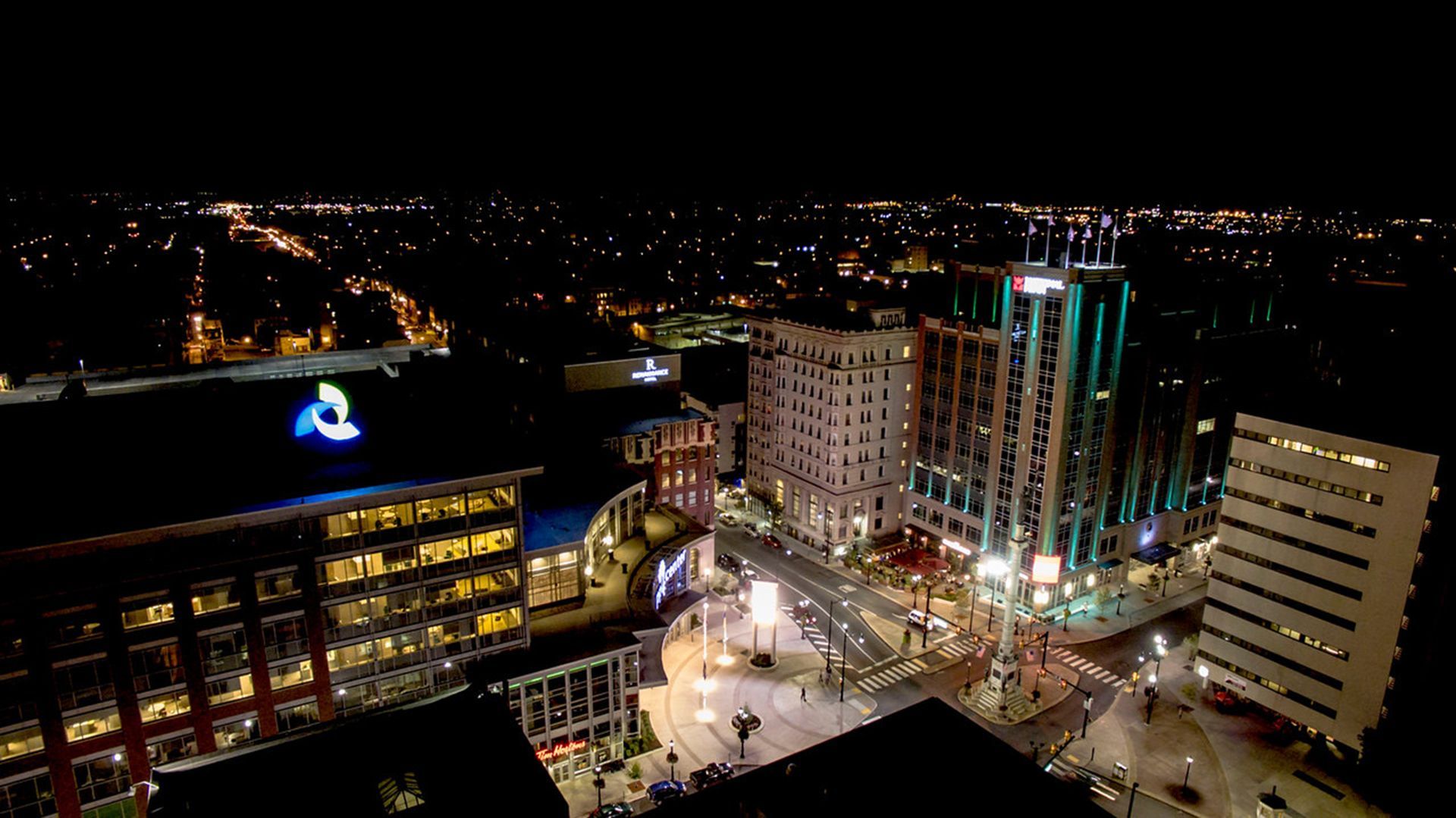 Nighttime cityscape with illuminated buildings, streets, and a lit plaza from above