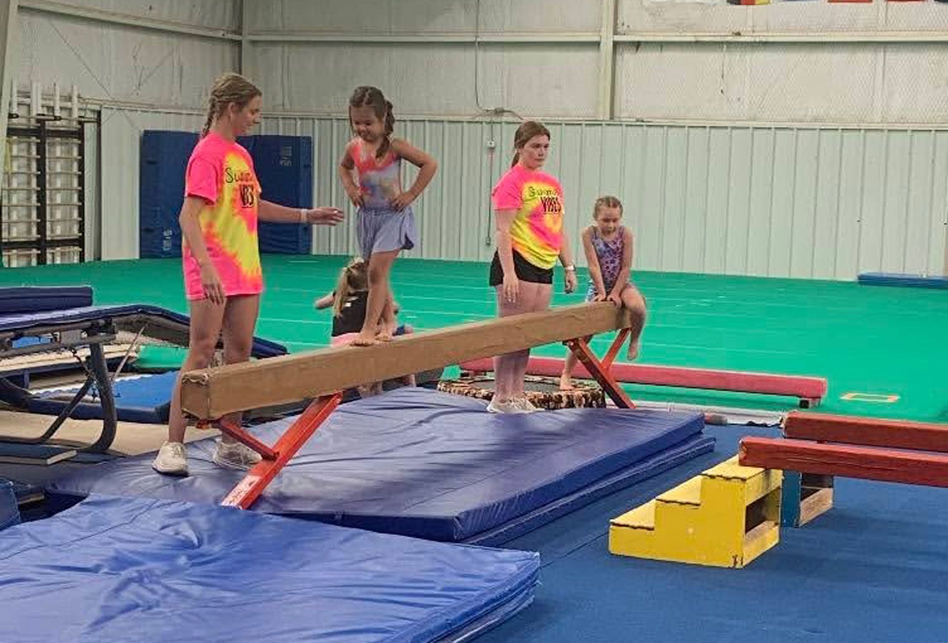 Three children balancing on a wooden beam in a gymnastics gym with mats and equipment nearby.