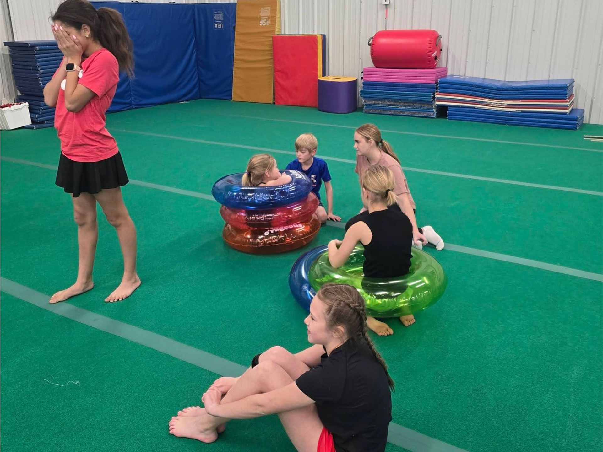 Children playing in colorful exercise rings on a gym floor, with one girl standing nearby and mats stacked in back.