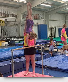 A woman is helping a young girl do a handstand on a parallel bars in a gym.