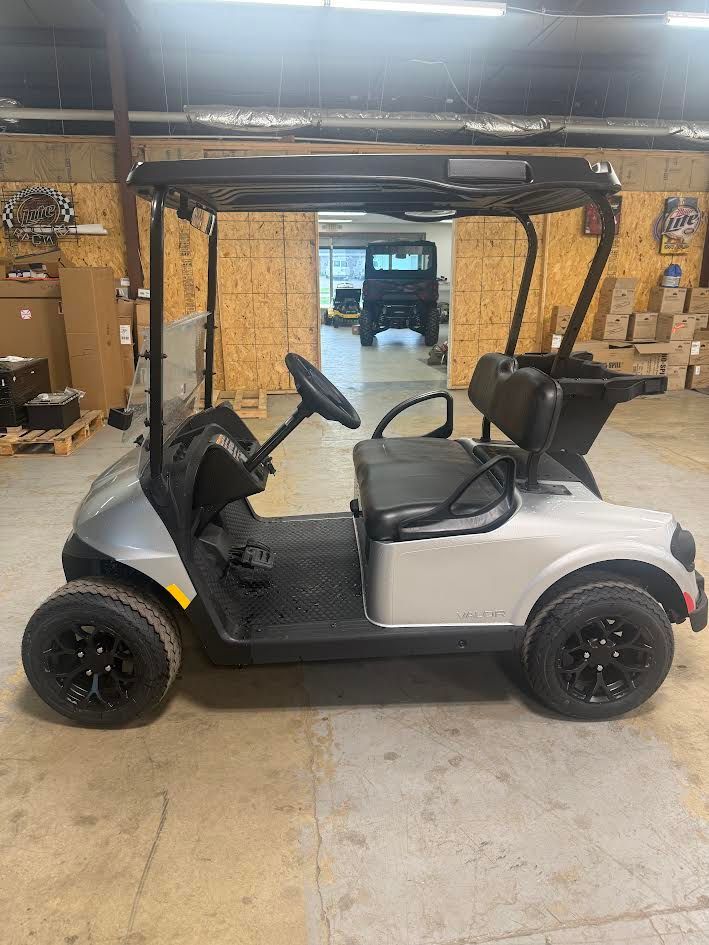 Silver golf cart parked inside a garage with a black roof and seats, viewed from the side.