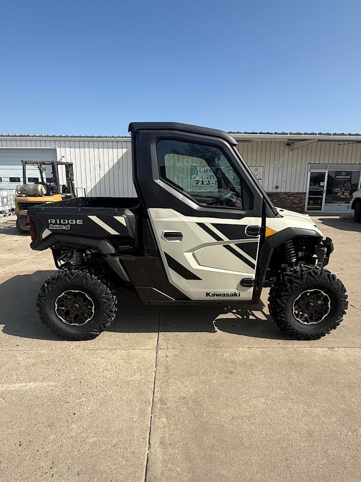 Side view of a black and beige Kawasaki UTV parked on a concrete lot in front of a building.