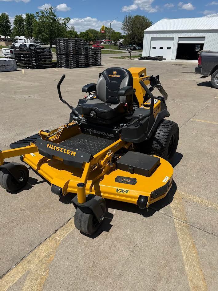 Yellow and black zero-turn mower parked on a concrete lot near a garage and stacked tires.