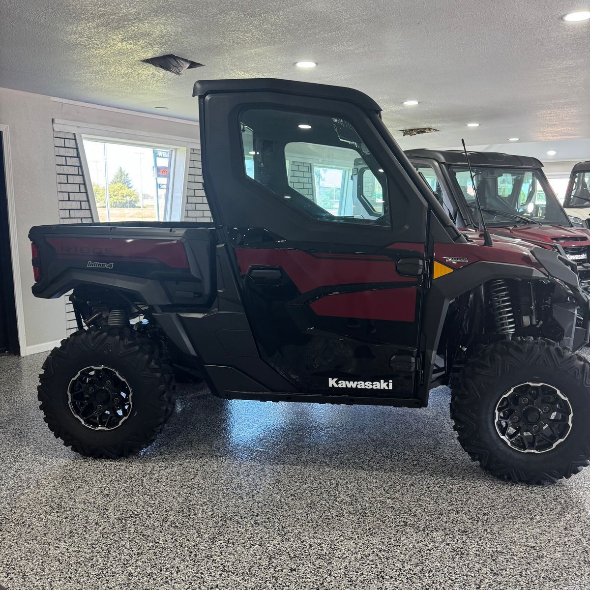 Red and black Kawasaki side-by-side utility vehicle displayed indoors on a showroom floor.
