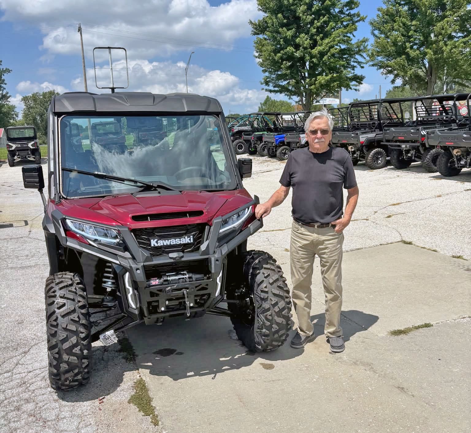 Man standing beside a red Kawasaki off-road vehicle in a parking lot with other ATVs in the background