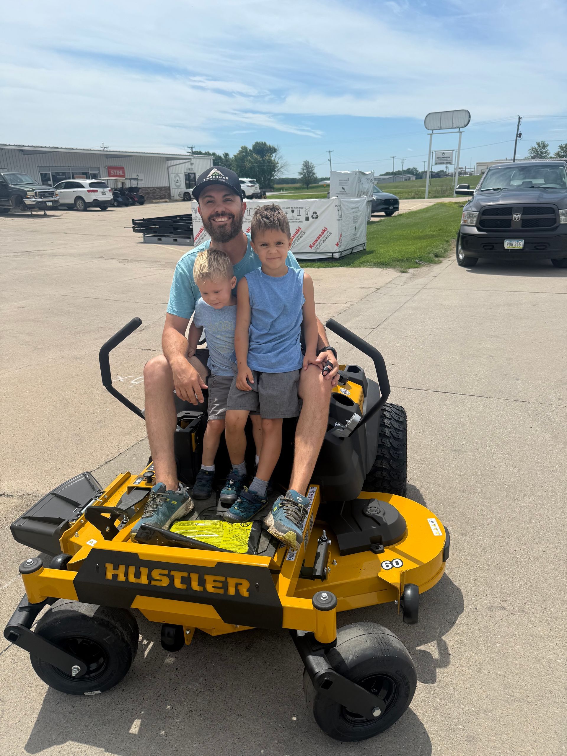 Man and two children on a yellow Hustler lawn mower in a parking lot