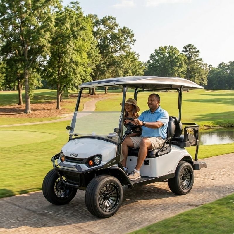 Two people driving a white golf cart on a paved path beside a green pond in a park