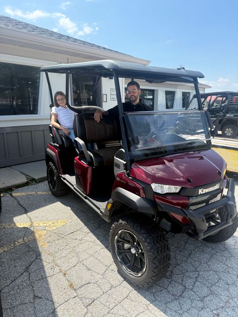 Two people ride in a red-and-black golf cart parked on a gravel lot beside a building.