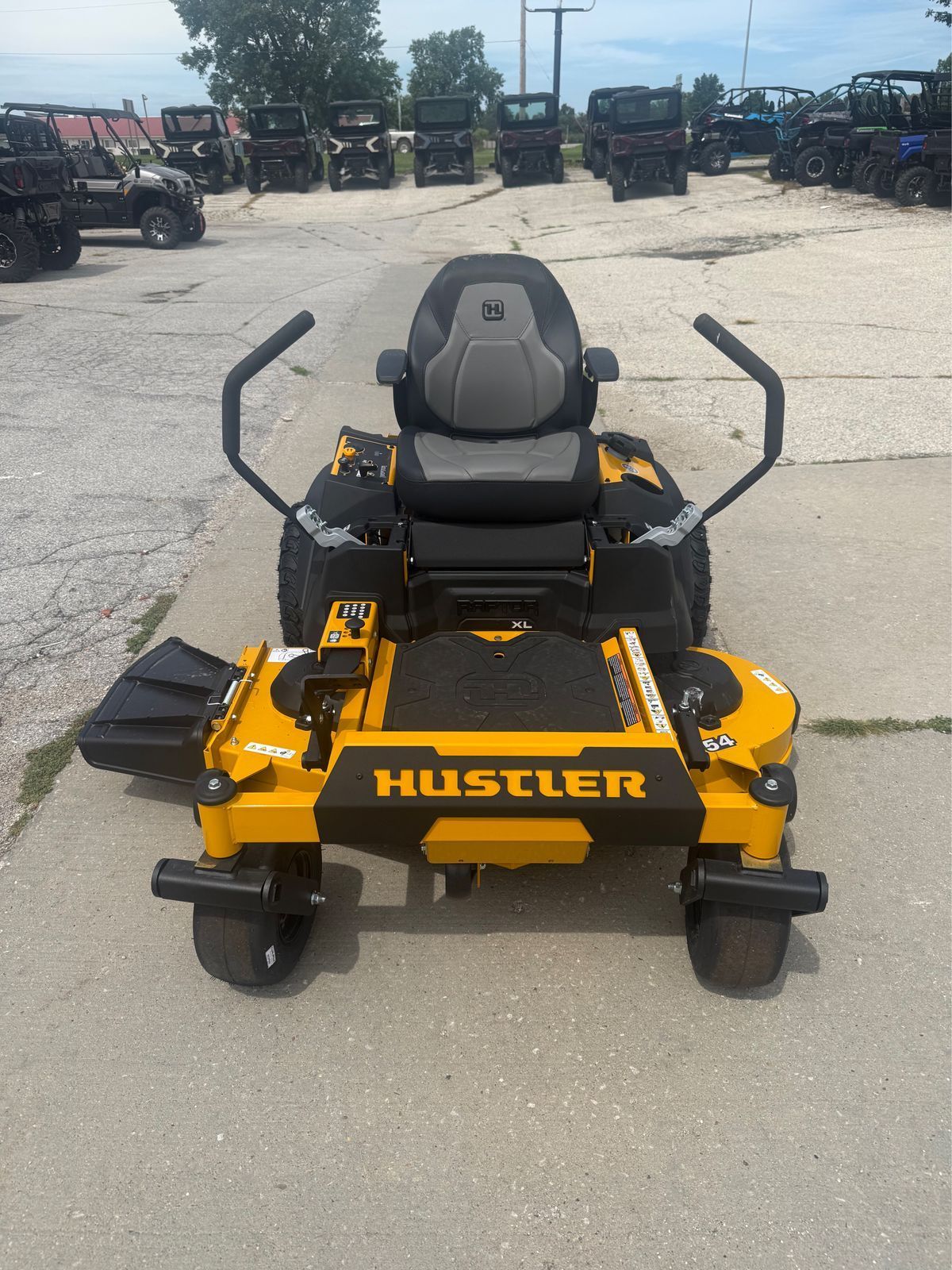 Yellow Hustler zero-turn lawn mower parked on pavement in an equipment lot, front view.