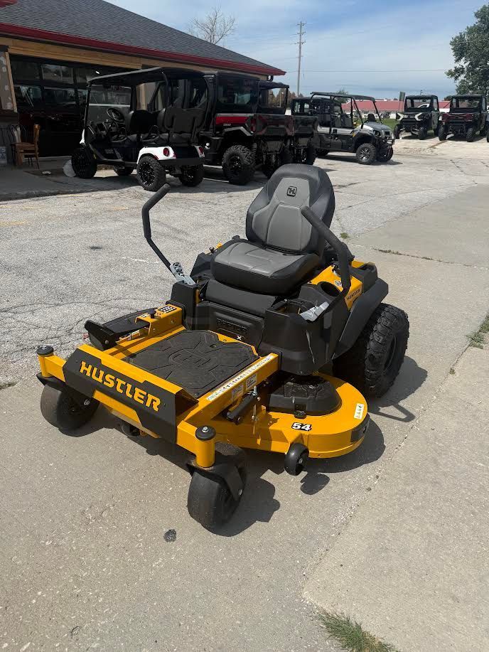 Yellow Hustler zero-turn lawn mower parked outdoors in a gravel lot near other utility vehicles.
