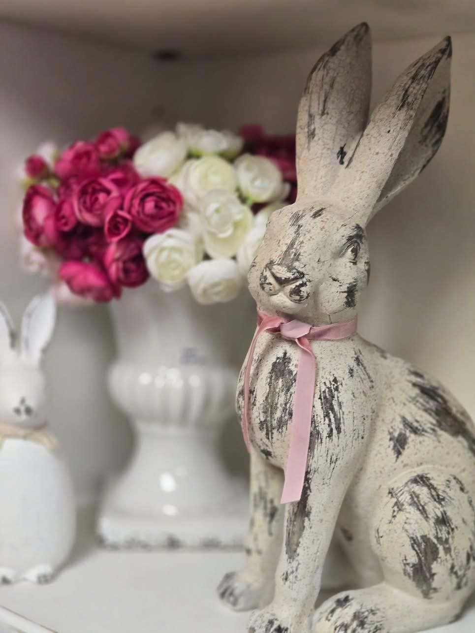 A white rabbit with a pink bow is sitting on a shelf next to a vase of flowers