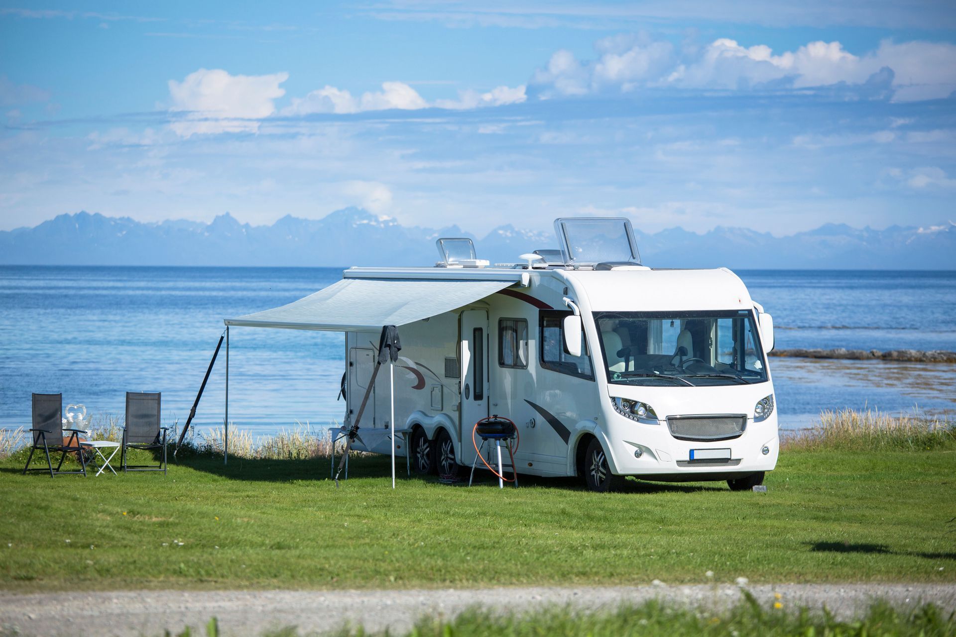 White camper van parked on grass by a calm blue lake under a cloudy sky, with awning extended.