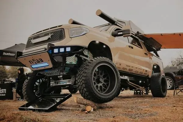 Beige off-road pickup truck lifted on large tires in a dirt race setting.