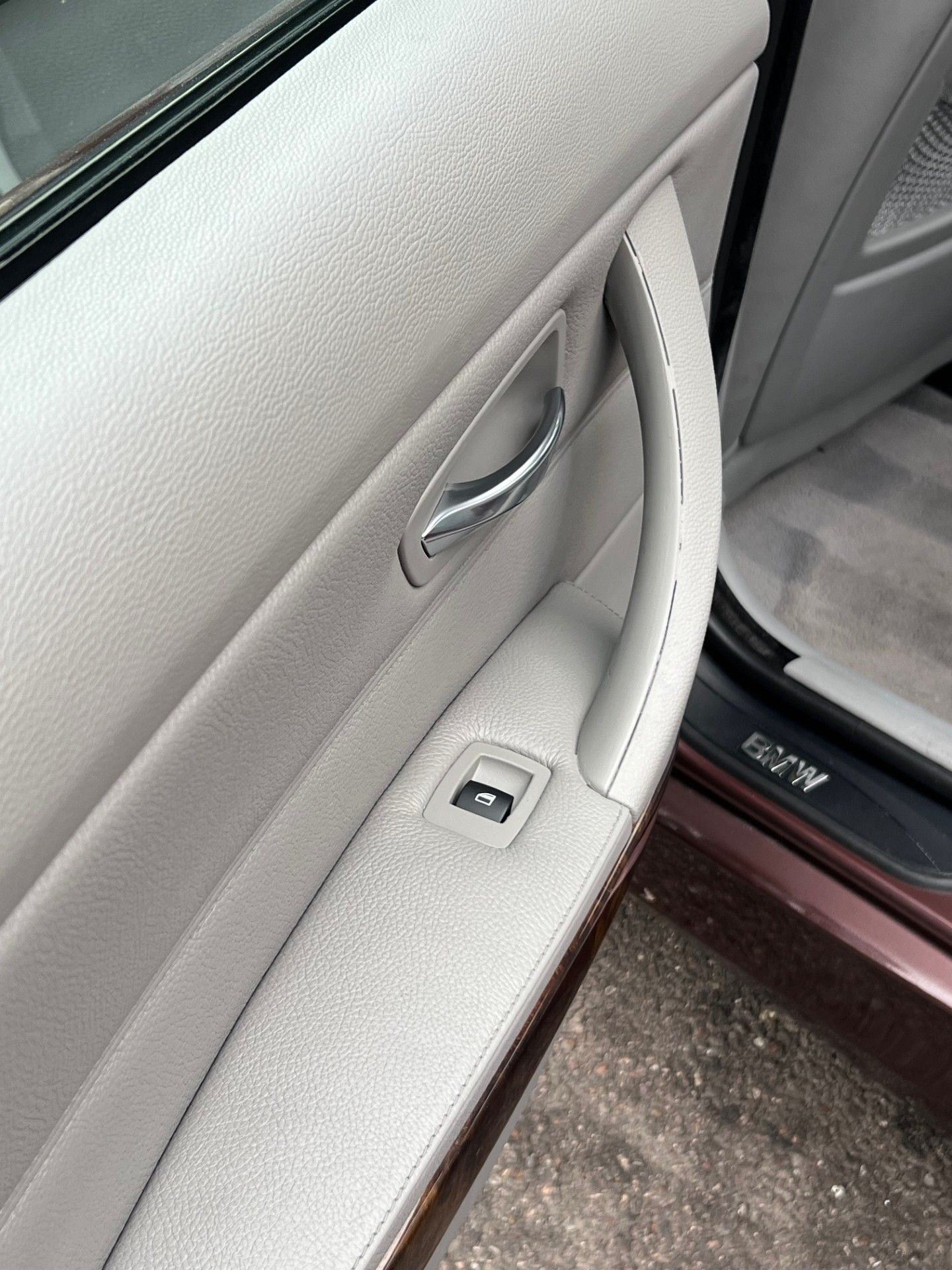 Interior of a light grey BMW vehicle door showing the door handle, armrest, and window control switch.