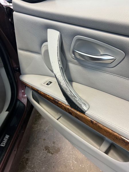 A BMW car door interior showing a damaged, peeling silver door grab handle against a light gray panel and wood trim.