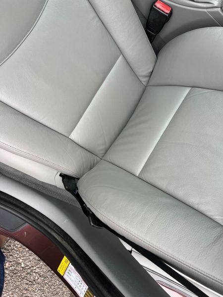 A light gray leather car seat showing a section of the seat cushion and backrest, with a black seatbelt buckle attached.